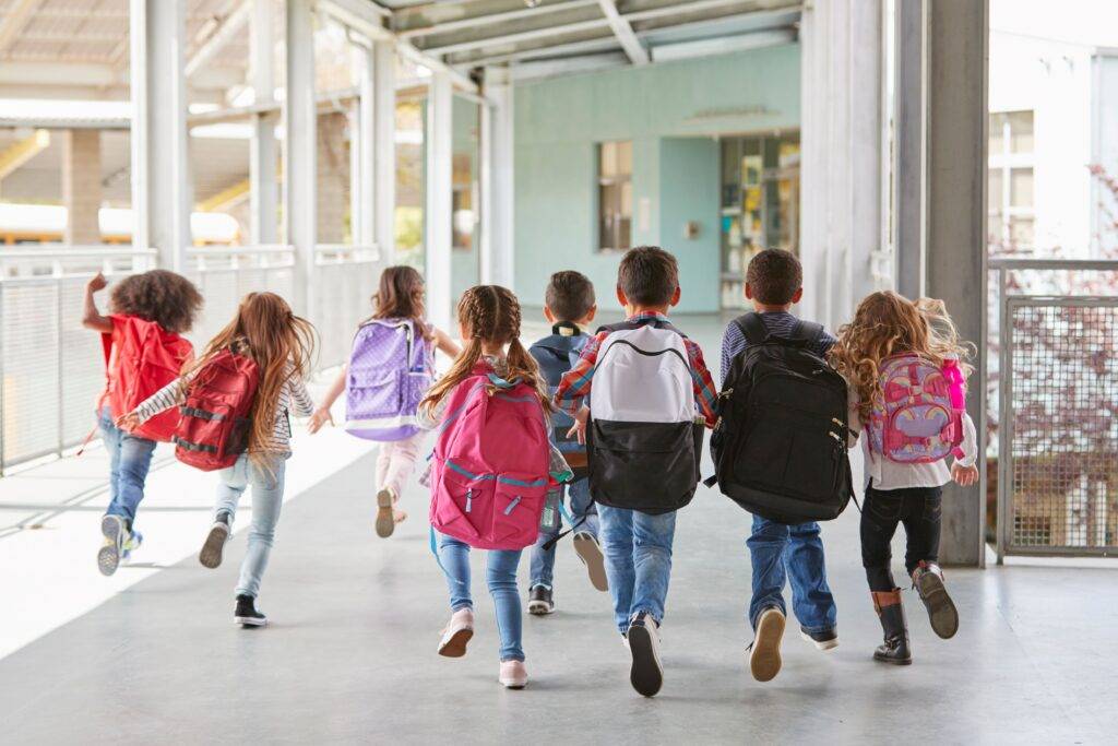 Gruppe von fröhlichen Kindern unterschiedlicher Ethnien rennt mit Rucksäcken durch einen hellen Flur in einer privaten Grundschule in Berlin, voller Energie und Begeisterung
