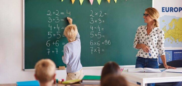 Junge Schülerin löst Mathematikaufgaben an der Tafel, während eine Lehrerin zuschaut, in einem Klassenzimmer einer privaten Grundschule in Berlin, dekoriert mit bunten Wimpeln und einer Europakarte im Hintergrund.