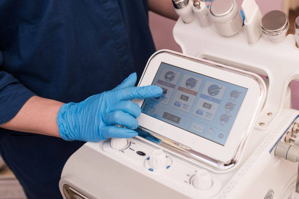 Kosmetikerin mit blauen Handschuhen bedient das Hydrafacial Gerät über ein Touchscreen-Display zur Vorbereitung der Hautbehandlung