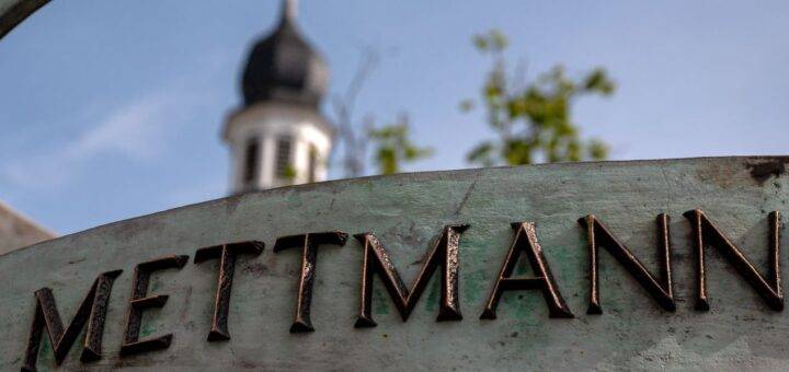 Bronze-Schild mit der Aufschrift "Mettmann" vor einem unscharfen Hintergrund, auf dem ein Kirchturm zu sehen ist – symbolisiert die Stadt Mettmann und ihren historischen Charme.