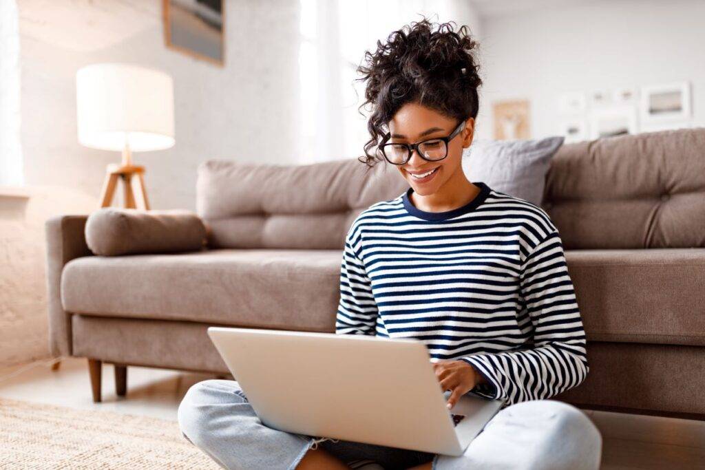 Junge Frau sitzt lächelnd auf dem Boden und arbeitet entspannt an ihrem Laptop im Wohnzimmer – symbolisiert die einfache Online-Kfz-Zulassung von Zuhause.