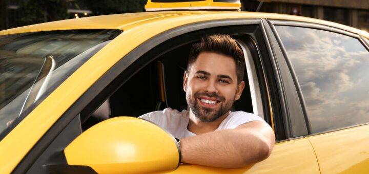 Taxifahrer Jobs: Junger Mann am Steuer eines gelben Taxis, lächelt freundlich aus dem Fenster